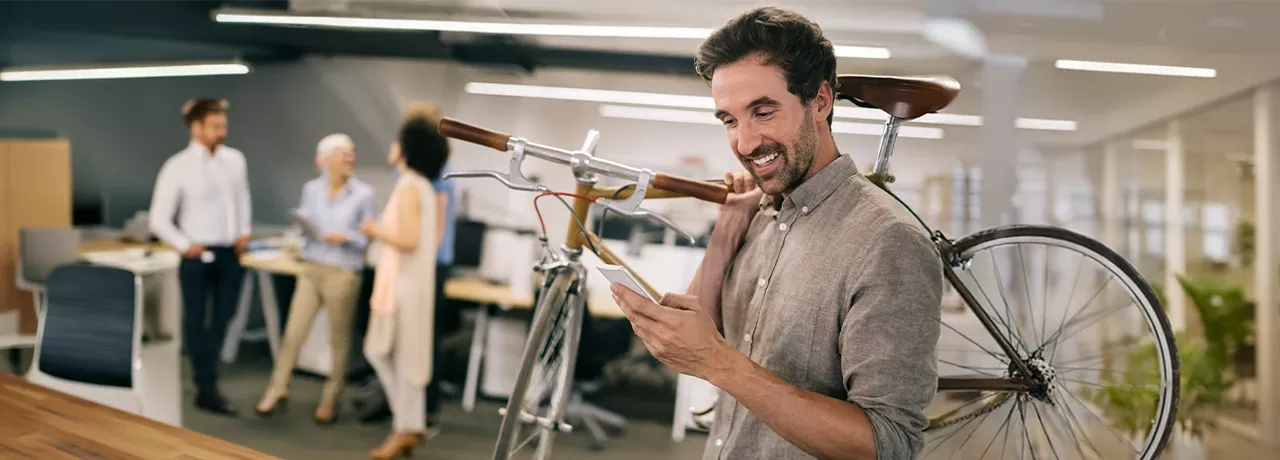 Mann im modernen Büro trägt sein Fahrrad auf der Schulter und schaut freudig auf sein Smartphone – Sinnbild für flexible Mobilität durch Bikeleasing. 