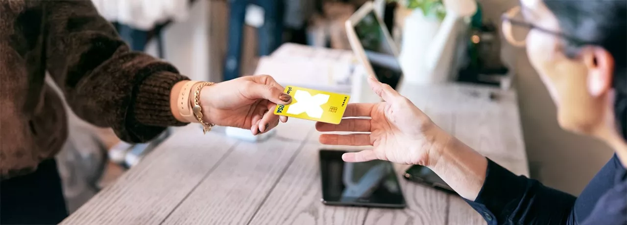 Hand mit braunem Pullover übergibt Pluxee Karte mit weißem Kreuz an andere Hand über Holztisch mit Tablet. 
