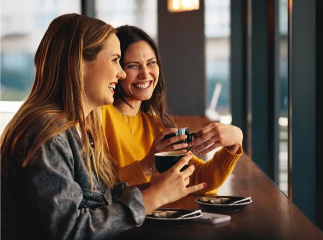 wei Frauen sitzen an einem Tisch mit Kaffeetassen und freuen sich über den Pluxee Benefit 10.000 Euro Bonus.