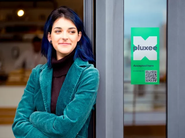 Eine Frau mit blauen Haaren steht vor einer Tür, auf der ein Pluxee-Aufkleber prangt.