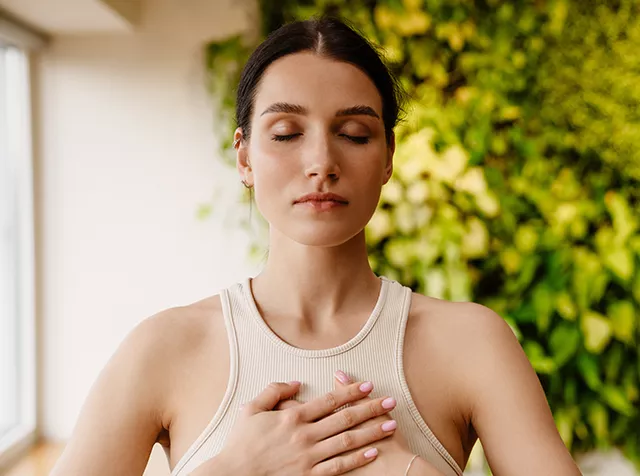 Frau in ärmellosem Oberteil hält Hände vor die Brust vor grünem Pflanzenhintergrund