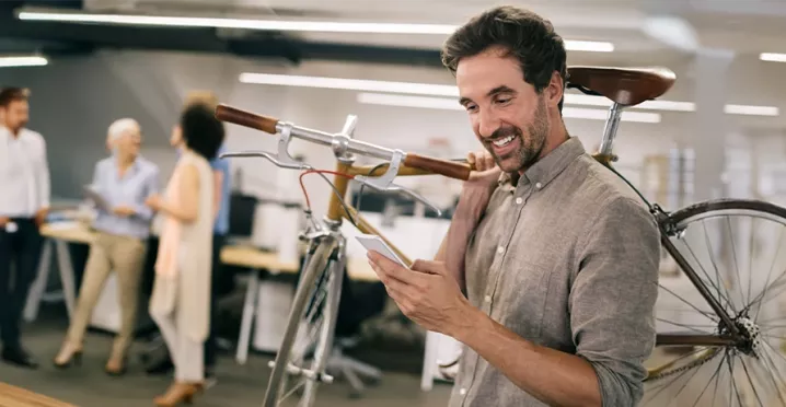 Mann im modernen Büro trägt sein Fahrrad auf der Schulter und schaut freudig auf sein Smartphone – Sinnbild für flexible Mobilität durch Bikeleasing.
