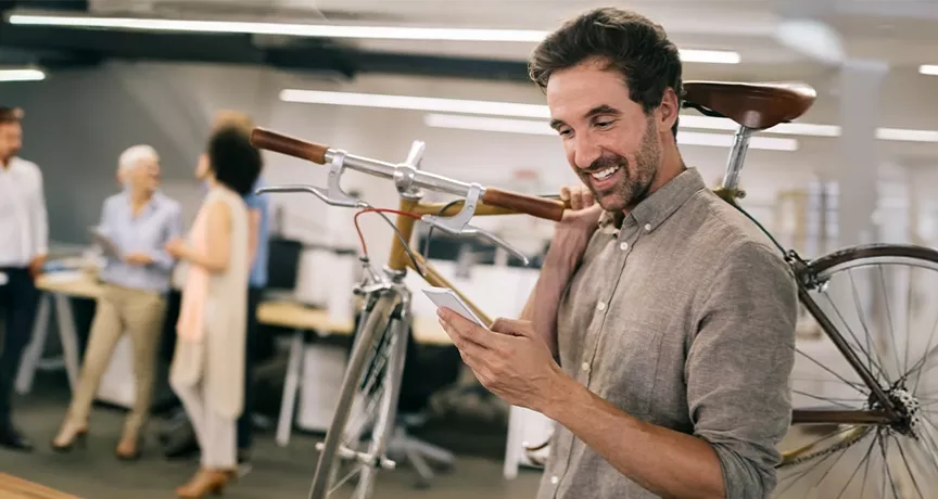Mann im modernen Büro trägt sein Fahrrad auf der Schulter und schaut freudig auf sein Smartphone – Sinnbild für flexible Mobilität durch Bikeleasing.