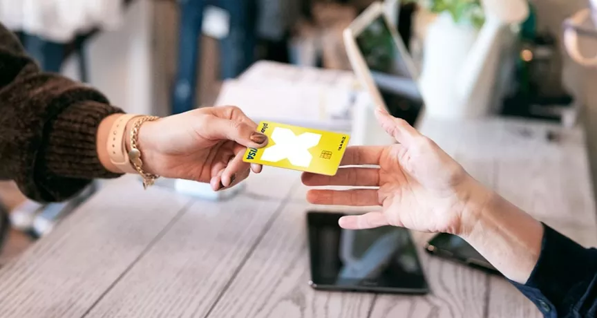 Hand mit braunem Pullover übergibt Pluxee Karte mit weißem Kreuz an andere Hand über Holztisch mit Tablet.