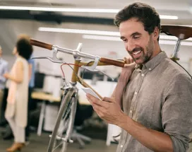 Mann im modernen Büro trägt sein Fahrrad auf der Schulter und schaut freudig auf sein Smartphone – Sinnbild für flexible Mobilität durch Bikeleasing.