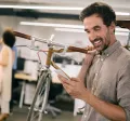 Mann im modernen Büro trägt sein Fahrrad auf der Schulter und schaut freudig auf sein Smartphone – Sinnbild für flexible Mobilität durch Bikeleasing.