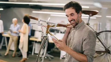 Mann im modernen Büro trägt sein Fahrrad auf der Schulter und schaut freudig auf sein Smartphone – Sinnbild für flexible Mobilität durch Bikeleasing.