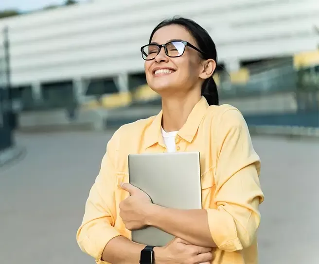 Lächelnde Frau mit Laptop im Arm im urbanen Umfeld – steht für berufliches Wohlbefinden, Zufriedenheit und moderne Arbeitswelt mit Pluxee.